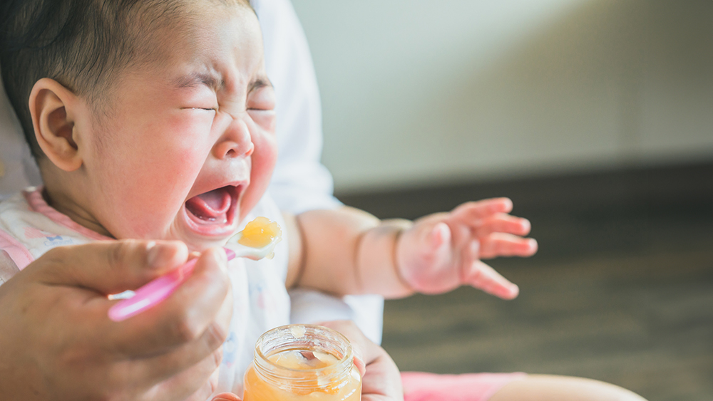 離乳食を食べさせられて泣いている赤ちゃんの写真